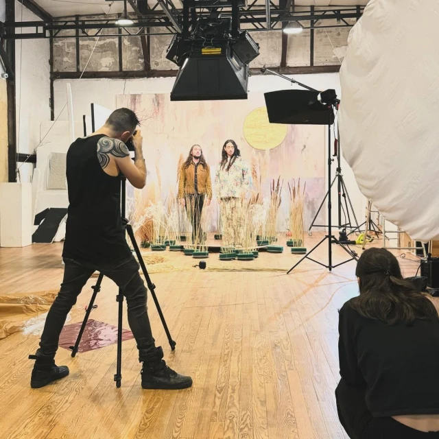 Photo studio shoot with two adults modeling patterned clothing, photographed by a man with tattoos on a tripod. Backstage crew and lighting.