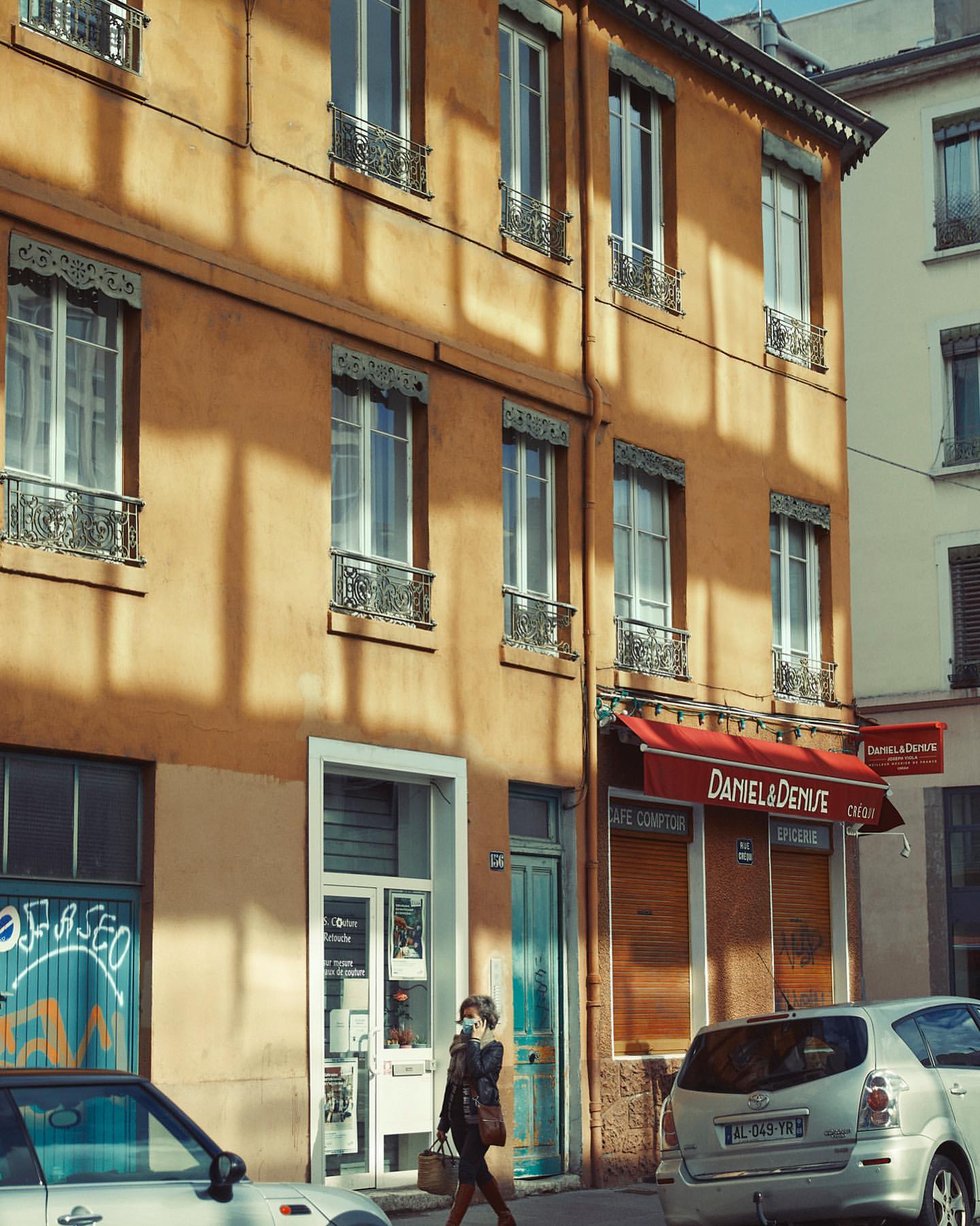 Woman in coat walks by tan buildings with awning-covered shop. Cars parked along street. City streetscape with people and architecture.