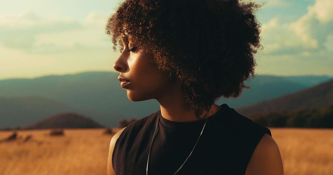 A young Black woman gazes pensively across a golden field, framed by rolling hills under a soft, partly cloudy sky at golden hour.