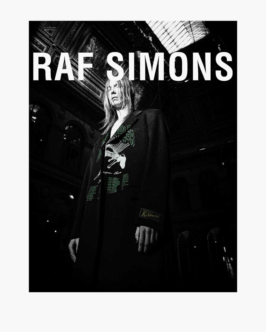 Model presents Raf Simons fashion, wearing patterned coat in a dimly lit interior with architectural details and bright overhead text.