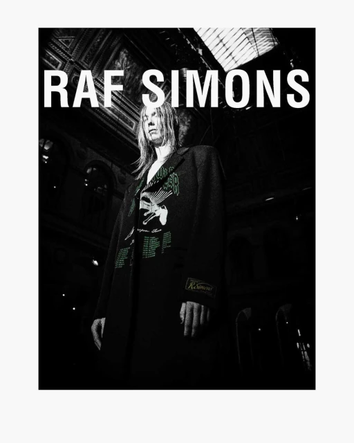 Model presents Raf Simons fashion, wearing patterned coat in a dimly lit interior with architectural details and bright overhead text.