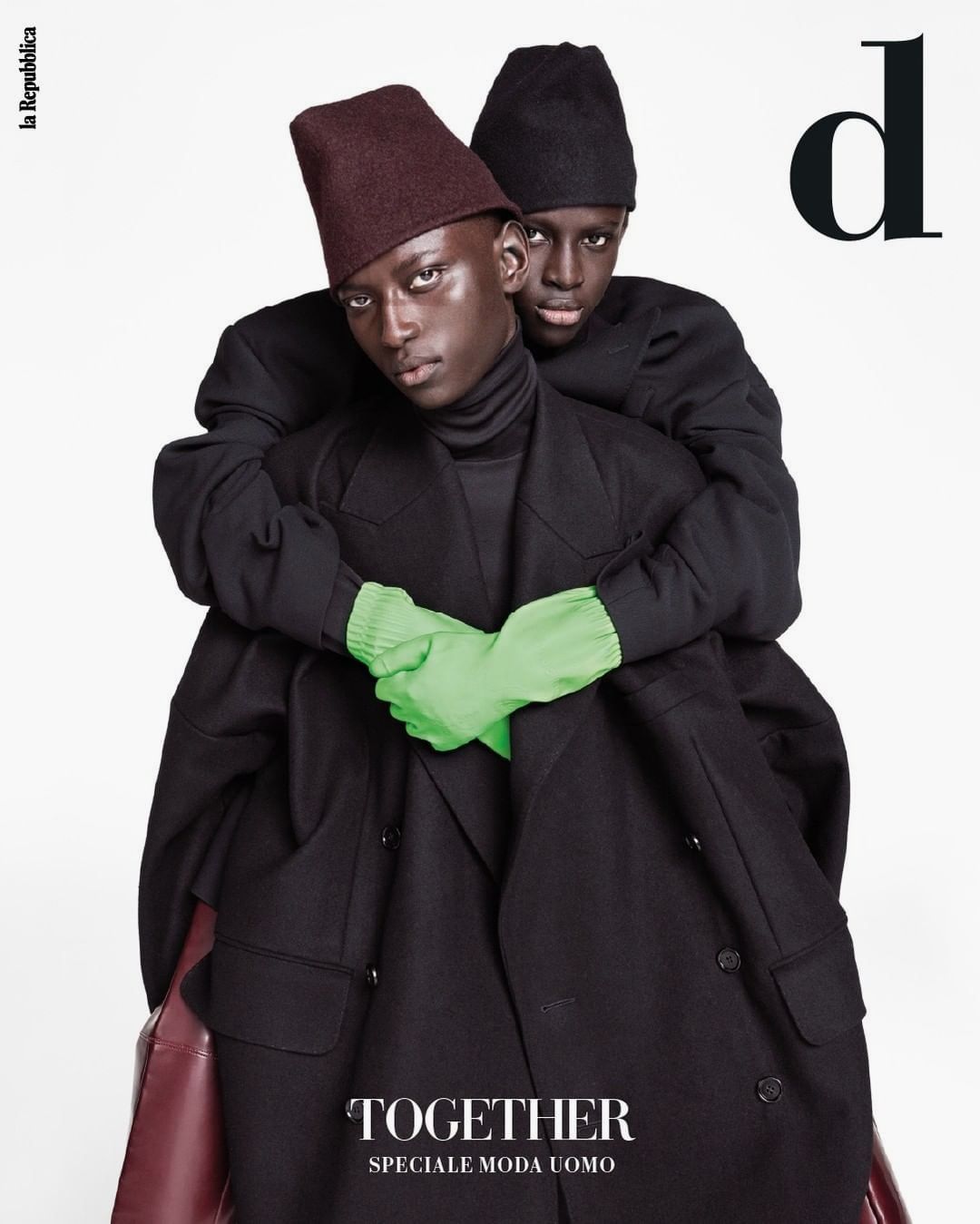 Two Black male fashion models embrace in black coats and hats, one in green gloves for a magazine cover against a white background.