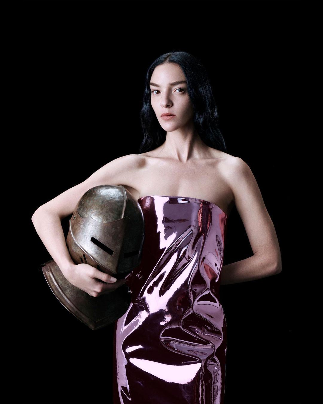 Woman in iridescent strapless dress holds helmet against black backdrop in studio. Beauty and fashion shot with dramatic lighting.