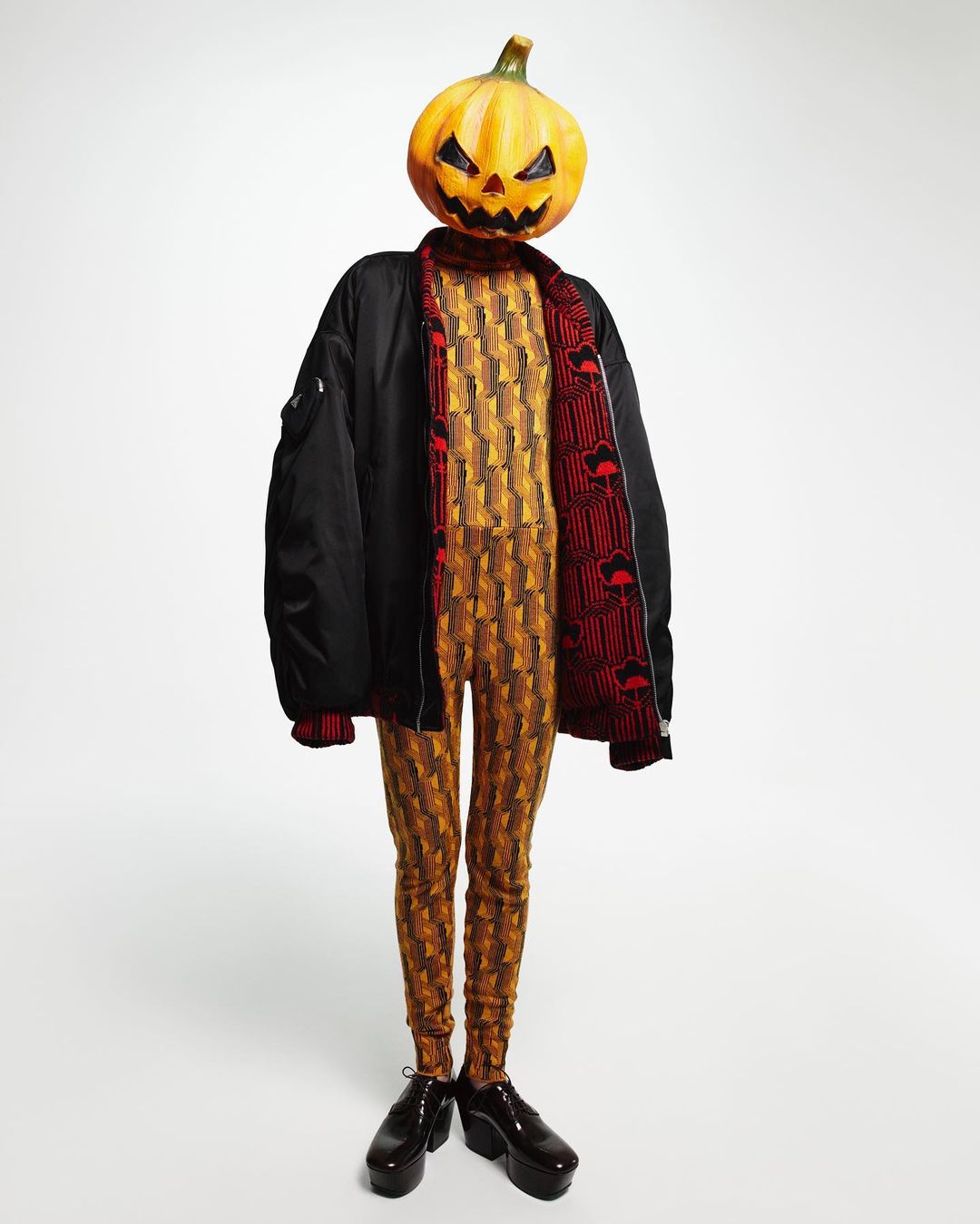 Person with a pumpkin head wearing a patterned jumpsuit and a black bomber jacket, against a white backdrop. Halloween-themed fashion.