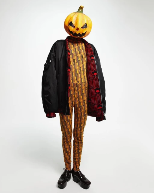 Person with a pumpkin head wearing a patterned jumpsuit and a black bomber jacket, against a white backdrop. Halloween-themed fashion.