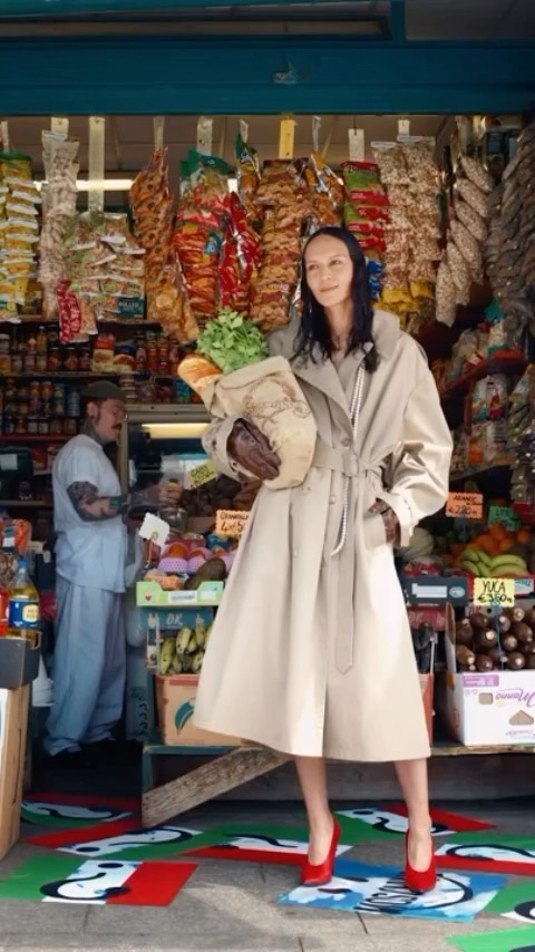 Woman in trench coat holding groceries stands in front of a store, with a person visible in the background.
