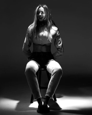 Black and white studio portrait of a woman seated on a stool wearing a button-down shirt and tall legwarmers.
