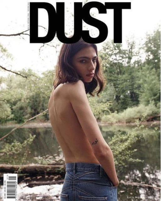 Magazine cover shot with a partially nude woman with dark hair, blue jeans, standing near a river and green trees, 'DUST' is the masthead.