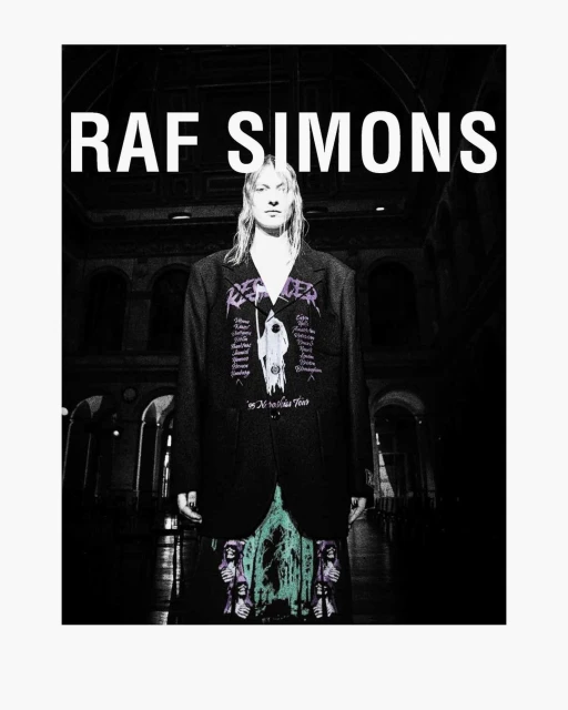 Fashion-forward editorial shot features a slender adult model in a black blazer and printed dress posing indoors against a dimly lit backdrop.