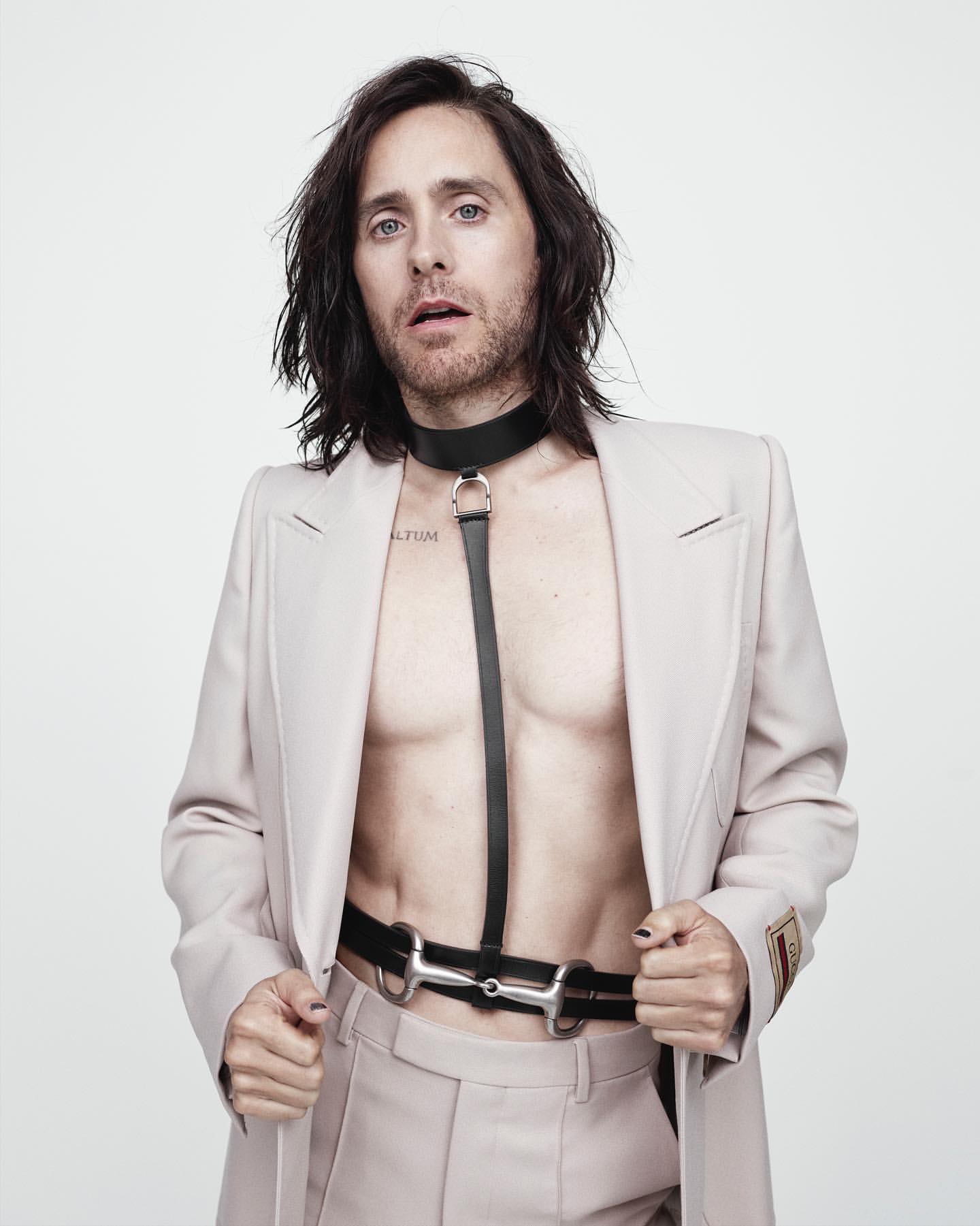 Jared Leto poses in a tan suit with a black leather harness against a white backdrop in a medium shot, fashion forward celebrity editorial.