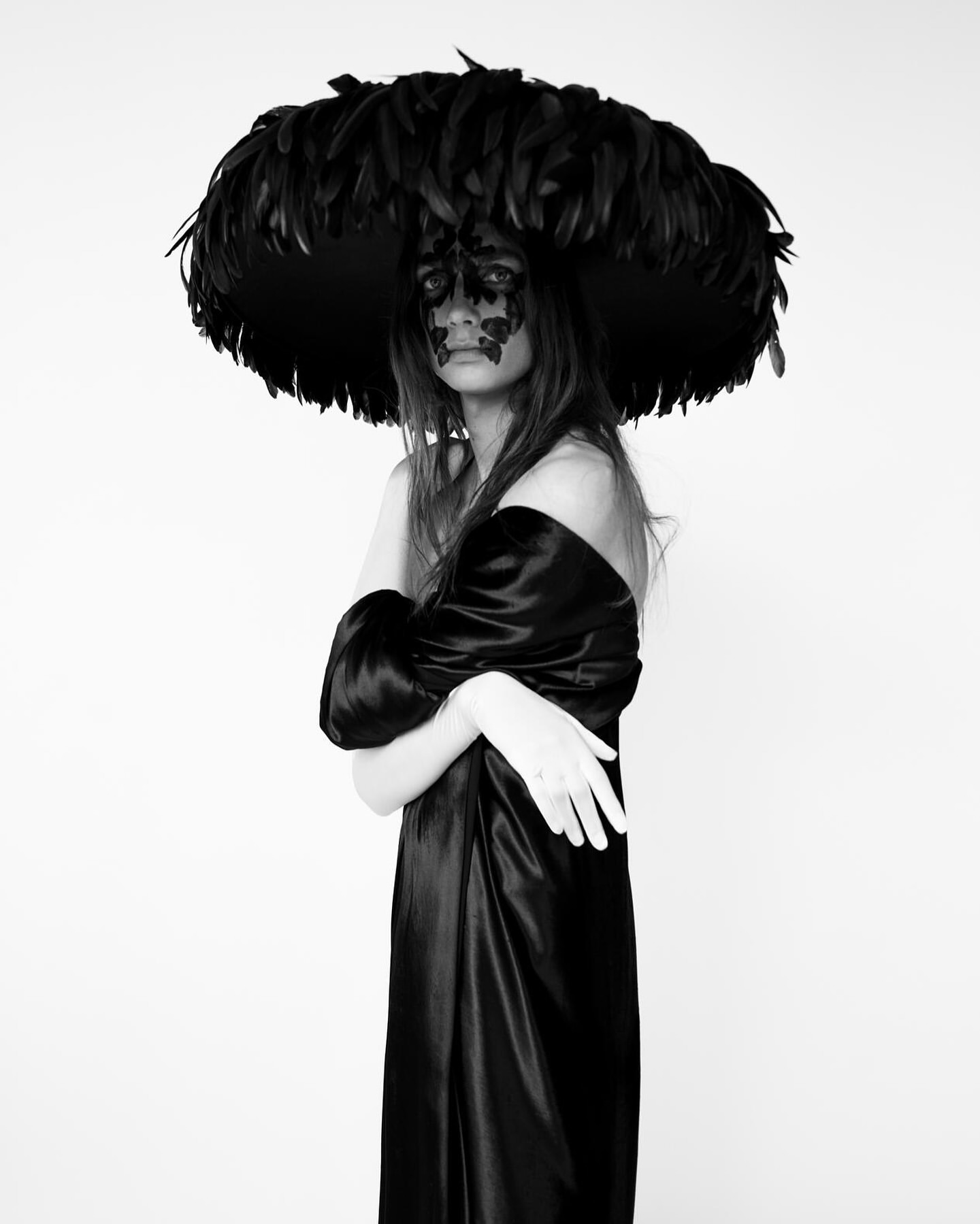 Woman wearing a large feathered hat and black dress in a studio. The image is black and white with a light background.