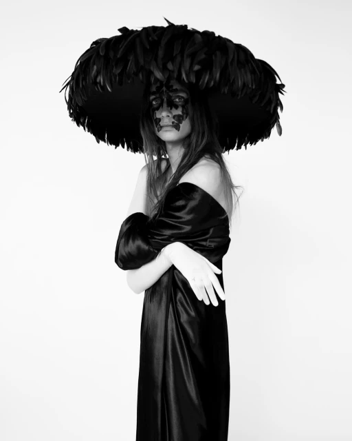 Woman wearing a large feathered hat and black dress in a studio. The image is black and white with a light background.