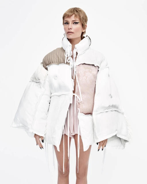 A model poses in a studio wearing an upcycled, white, deconstructed puffer jacket with neutral accents against a white background.