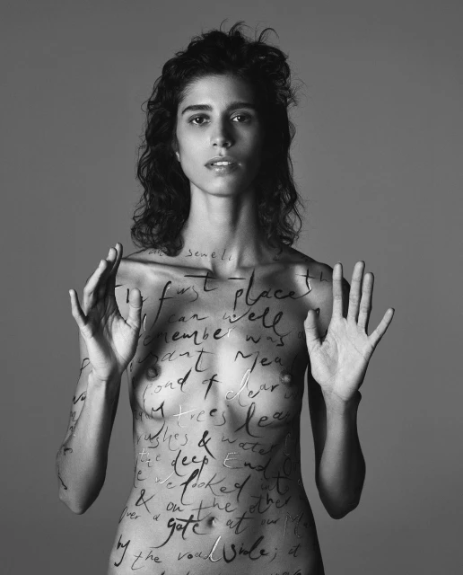 Monochrome portrait of a woman with handwritten text art covering her body, her hands raised, set against a grey backdrop.