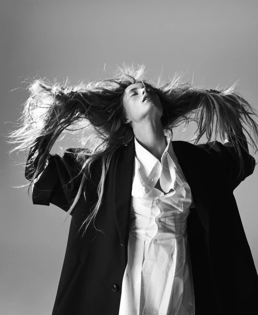 Monochrome portrait of a model with long hair tossed back, wearing a blazer and white top, against a neutral background in a studio shot.