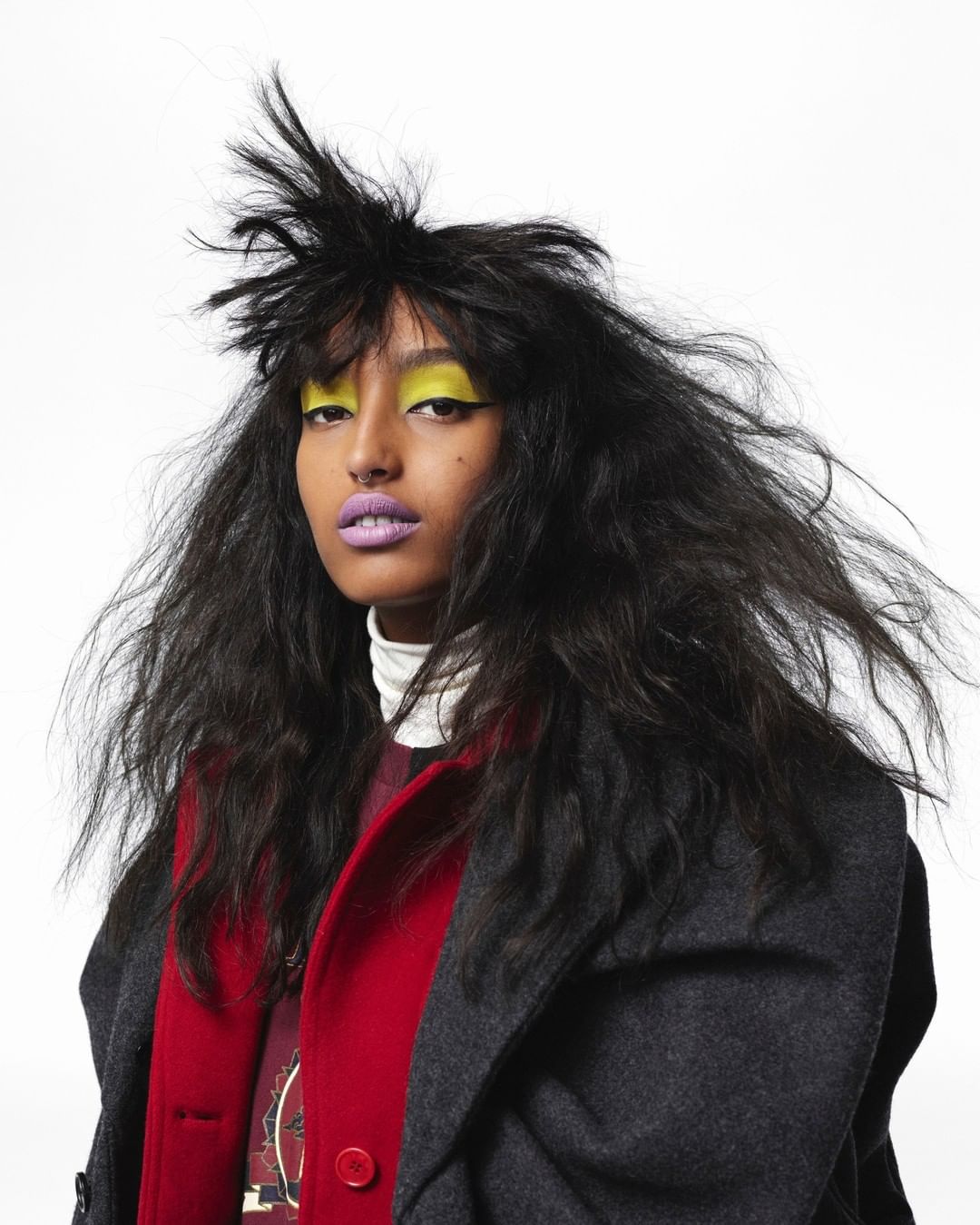 A striking portrait of a woman with dramatic makeup, including vibrant yellow eyeshadow and lilac lipstick, against a crisp white background.