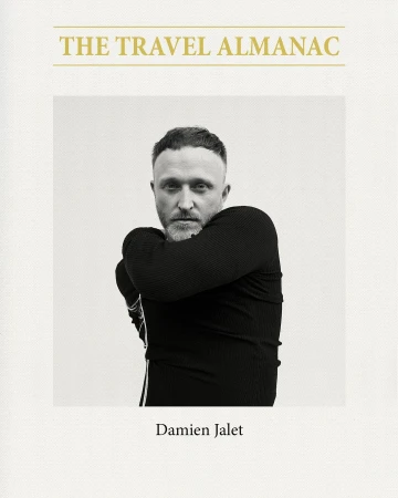 Black and white portrait of a man wearing a black top against a light background, on the cover of a book titled 'The Travel Almanac'.