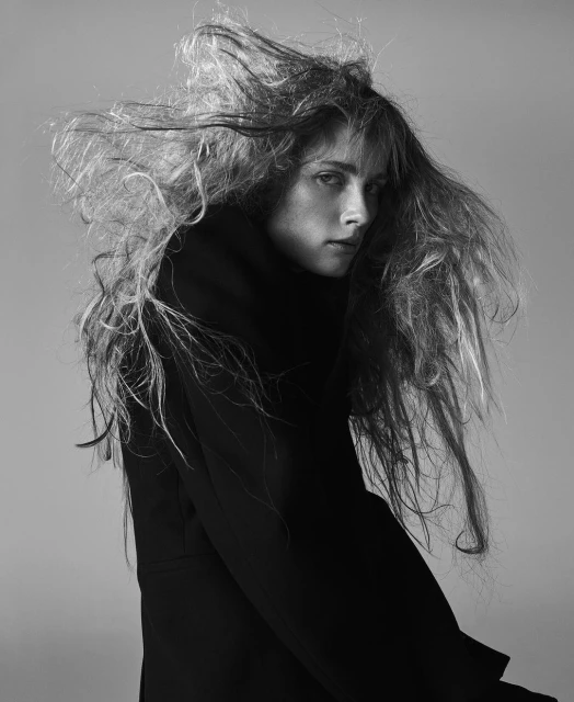 Black and white portrait of a female model with long hair, wearing a dark coat, against a grey background in a photo studio.