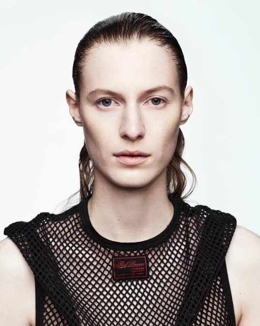 Headshot of a fair-skinned woman with wet hair wearing a black mesh top against a white background in a studio setting.