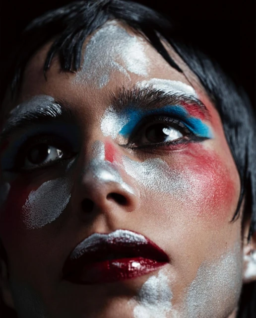 Dramatic beauty portrait: a person with silver, blue, and red artistic makeup on their face against a dark background.
