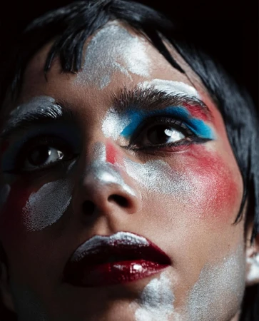 Dramatic beauty portrait: a person with silver, blue, and red artistic makeup on their face against a dark background.