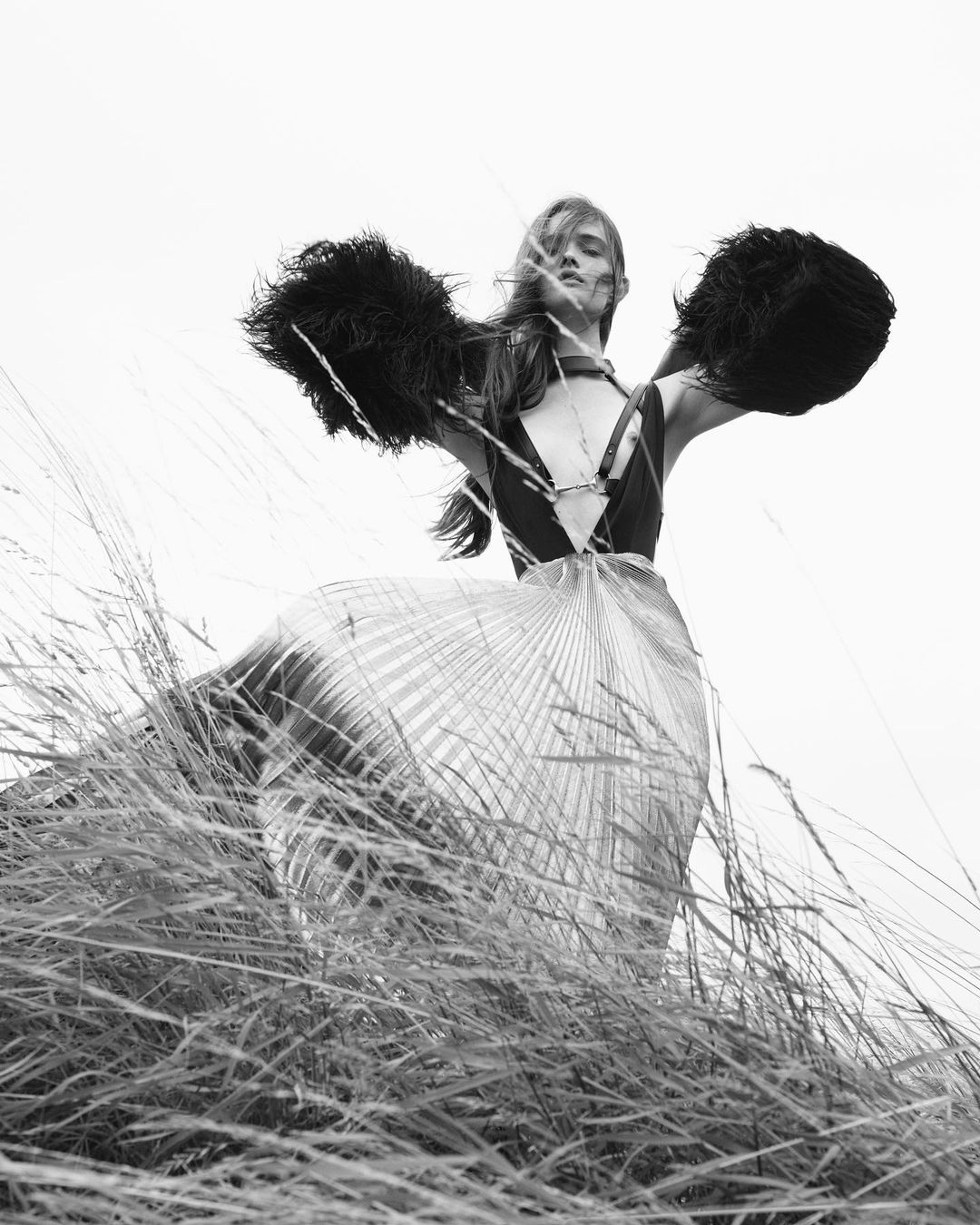 Black and white fashion shot of a woman in a flowing skirt and feathered shawl standing in a field of tall grass against a bright sky.