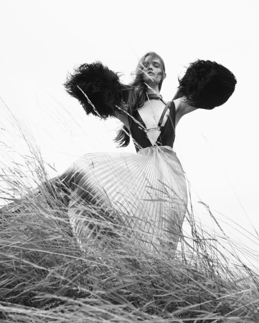 Black and white fashion shot of a woman in a flowing skirt and feathered shawl standing in a field of tall grass against a bright sky.