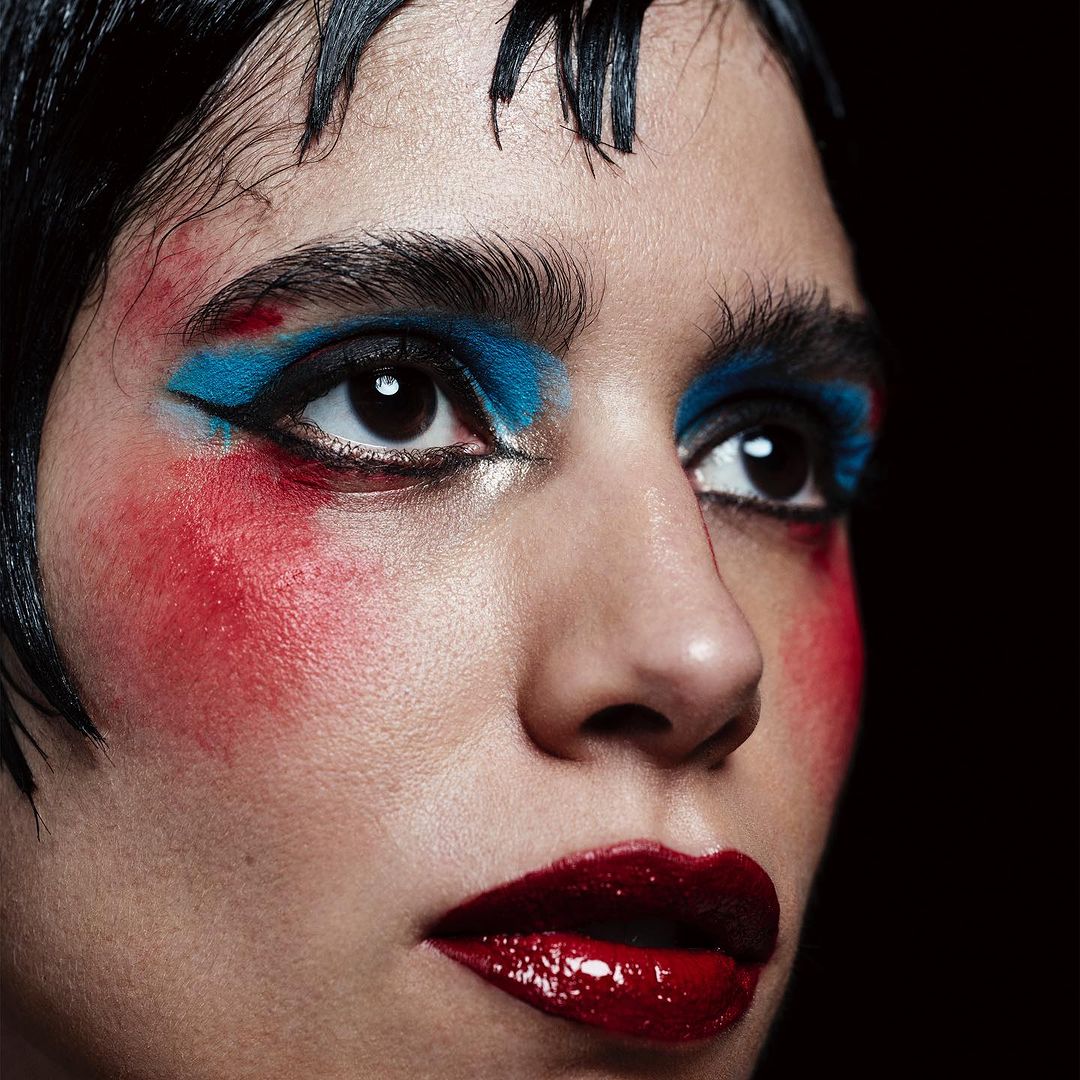 Close-up portrait of a woman with bold blue eyeshadow, red blush, glossy red lips, and dark wet-look hair against a black background.