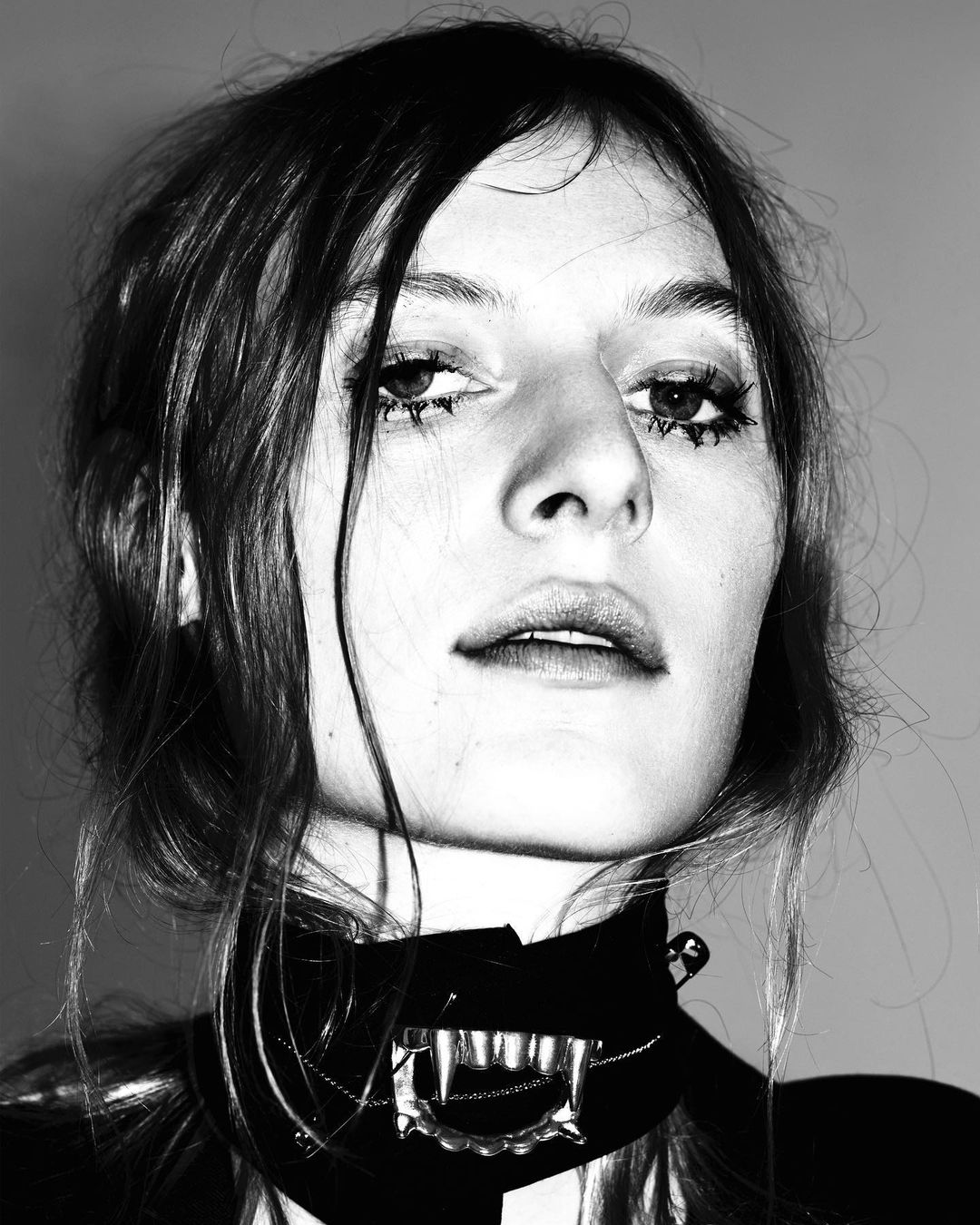 Black and white portrait of a woman with striking makeup, wearing a distinctive fang necklace, in a close-up studio setting.