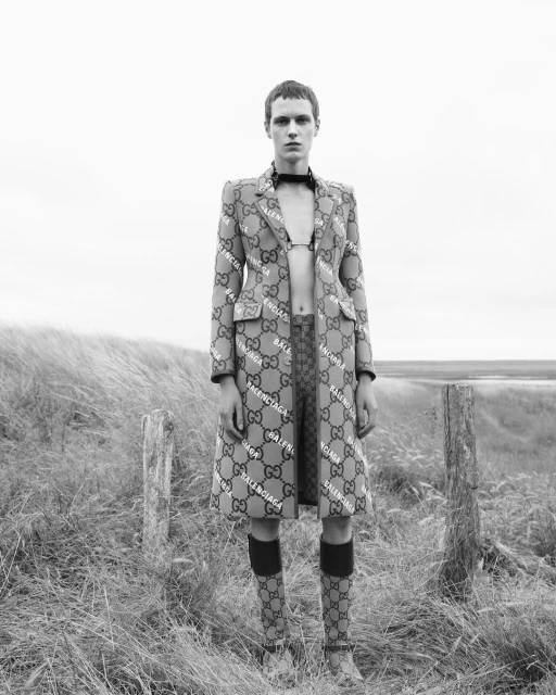Monochrome fashion shot: person wearing Balenciaga and Gucci logo coat, bralette, and socks stands outdoors in a grassy, neutral field.