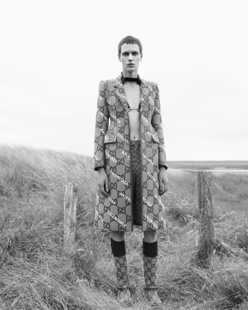 Monochrome fashion shot: person wearing Balenciaga and Gucci logo coat, bralette, and socks stands outdoors in a grassy, neutral field.