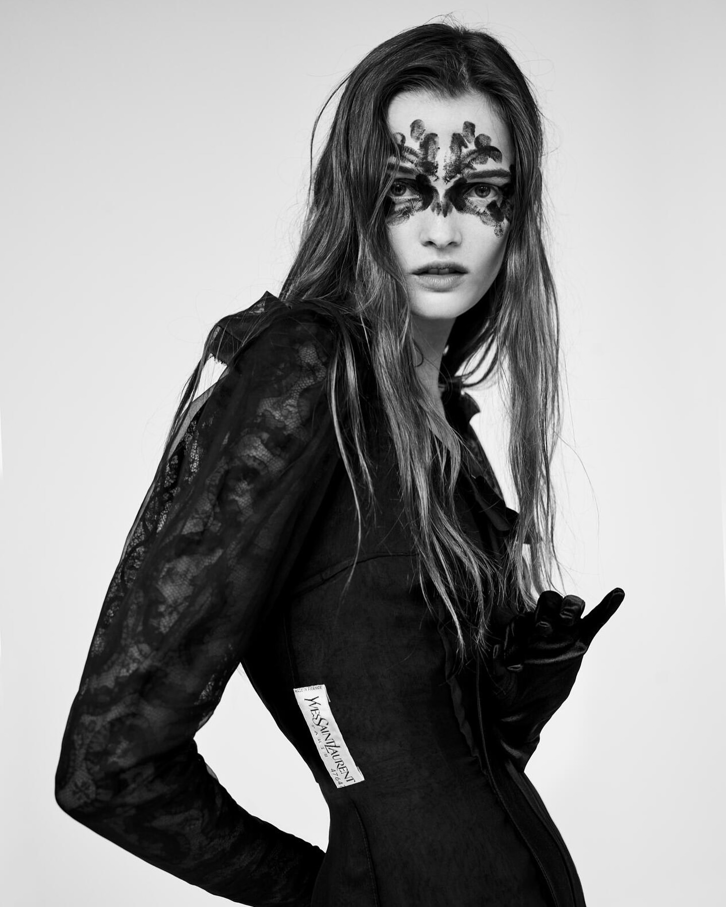 Fashion portrait of a female model with abstract black makeup, long brown hair, wearing a dark lace dress and gloves against a grey background.
