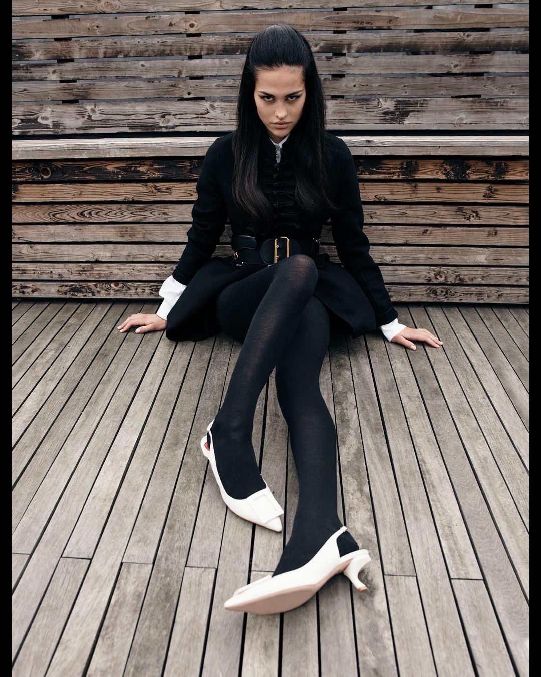 A woman in a black coat and tights sits on a wooden surface, showcasing white high-heeled shoes in an outdoor fashion shot.