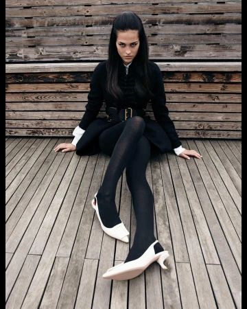 A woman in a black coat and tights sits on a wooden surface, showcasing white high-heeled shoes in an outdoor fashion shot.