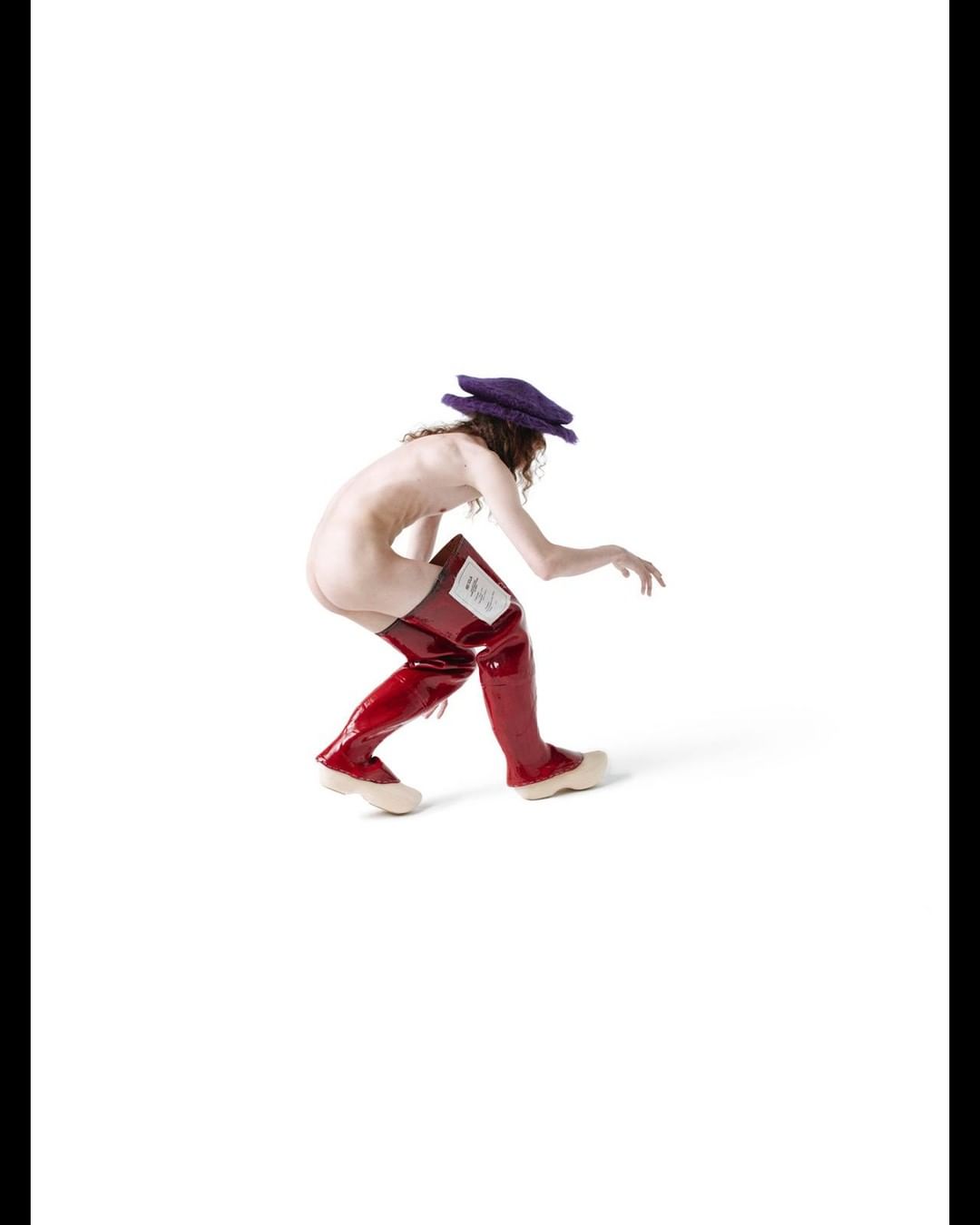 Artistic studio shot of a nude adult female wearing red thigh-high boots and a purple hat, striking a crouching pose in a bright white studio.