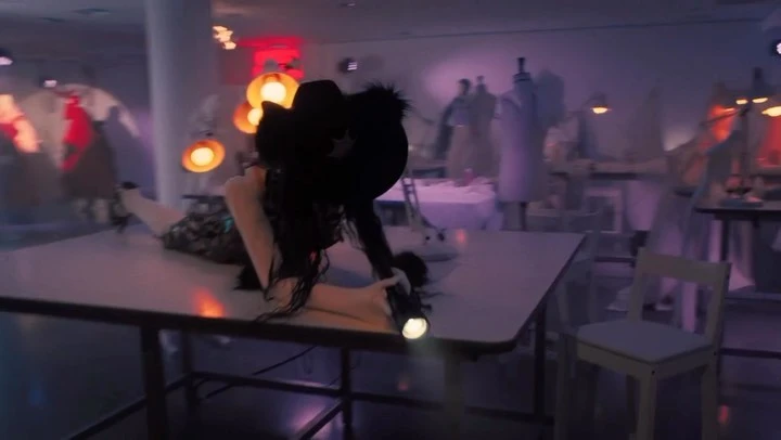 Woman in black hat poses on a table in a fashion design workshop with sewing mannequins and bright orange lighting fixtures in the background.