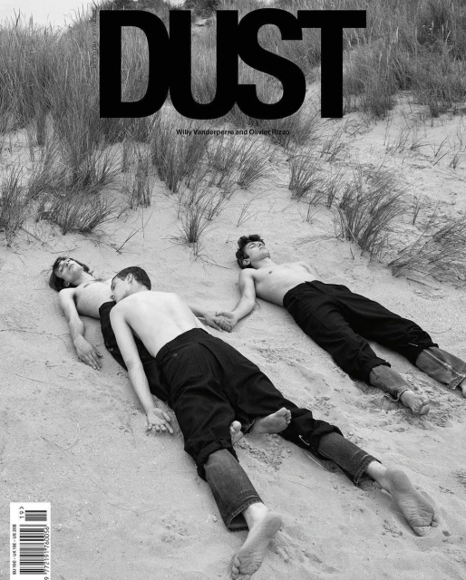Black-and-white fashion editorial cover featuring three shirtless young men holding hands on a sandy beach with grassy dunes.