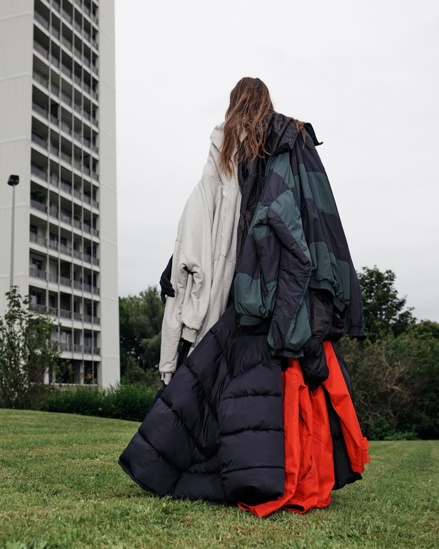 Woman stands on green grass wearing multiple puffer jackets in an outdoor setting with a tall building in the background on an overcast day.