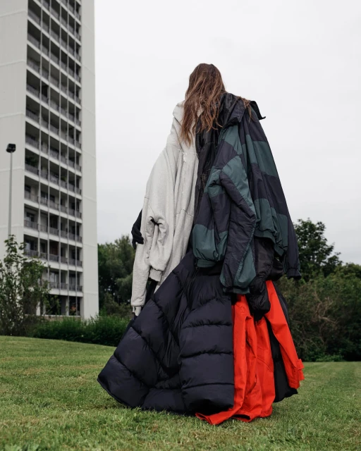 Woman stands on green grass wearing multiple puffer jackets in an outdoor setting with a tall building in the background on an overcast day.