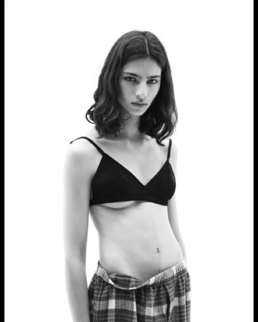 Monochrome portrait of a slender woman with medium-length wavy dark hair, wearing a bra and pajama pants against a bright white backdrop.