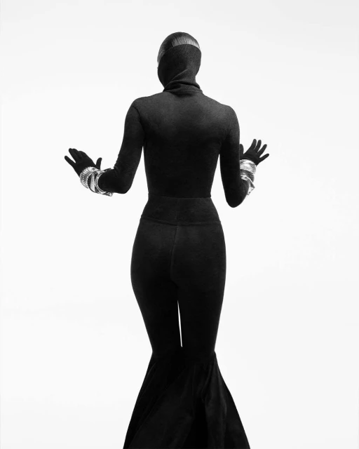 Back view of a slender, hooded woman in dark clothing with silver bracelets against a bright white background in a studio setting.