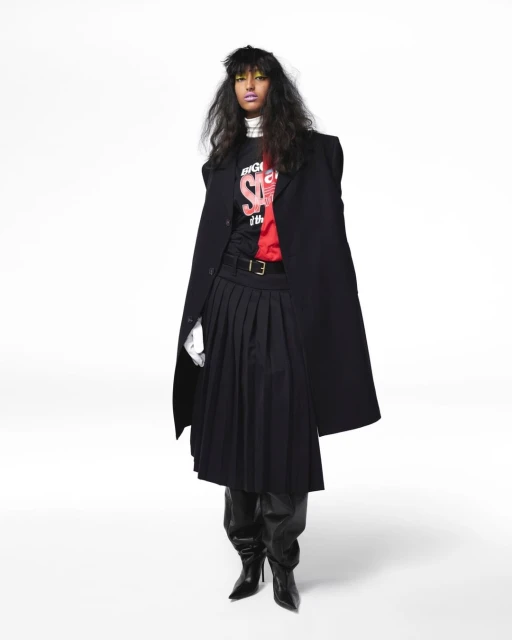 Fashionable woman with dark hair, wearing a long black coat, pleated skirt, t-shirt with text, and black boots, posing indoors.