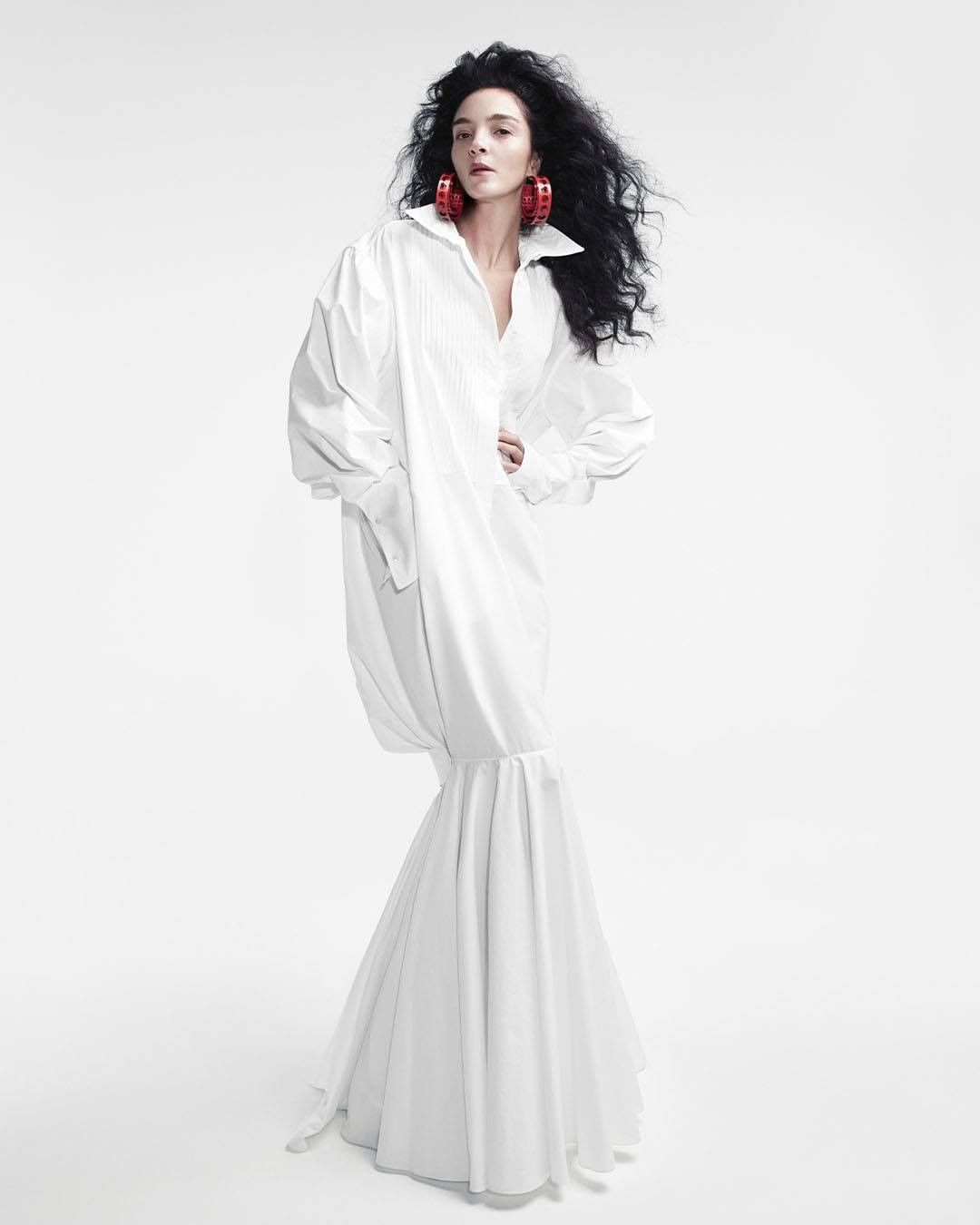 Fashion photo of a woman with dark curly hair in a long, oversized white shirt dress, posing against a bright white backdrop in a studio setting.