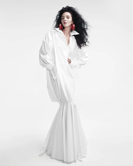Fashion photo of a woman with dark curly hair in a long, oversized white shirt dress, posing against a bright white backdrop in a studio setting.