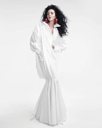 Fashion photo of a woman with dark curly hair in a long, oversized white shirt dress, posing against a bright white backdrop in a studio setting.