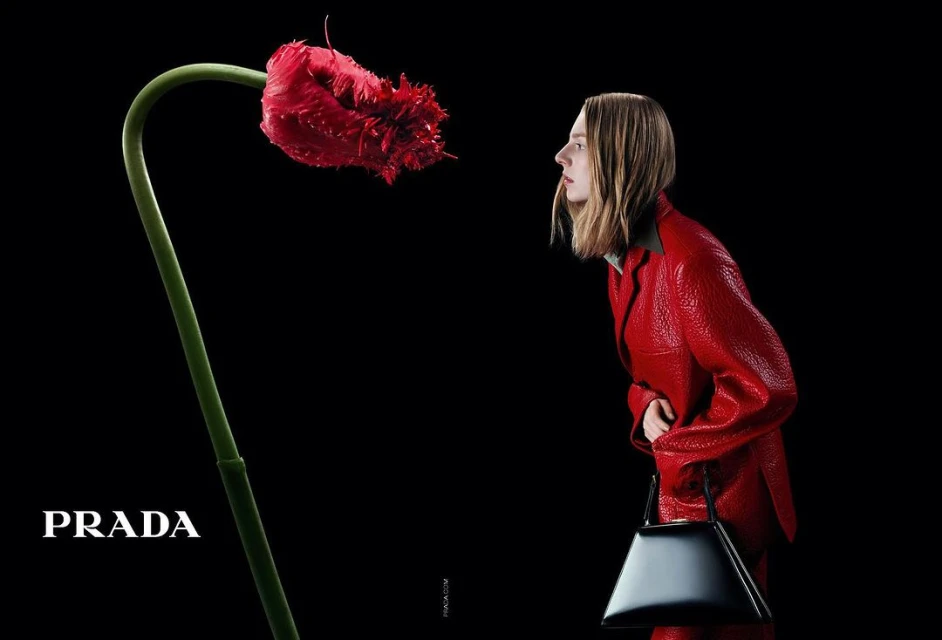 Woman in red suit gazes at a vibrant red flower against black background. Prada logo visible. Fashion advertising or editorial.