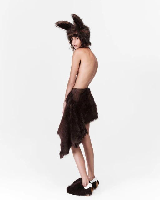 Woman wearing fur hat with ears, fur skirt, and slippers, standing in a white studio. Fashion or costume concept. Neutral expression.