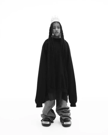 Young person with braided hair wearing an oversized black hoodie, loose gray pants, platform shoes, and a silver crown against a white background.