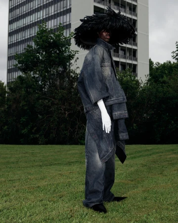 Model in oversized denim outfit wearing large feathered hat and white gloves standing on grass before a tall building