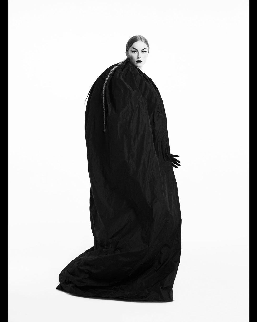 Woman wearing an oversized, draped black garment in a fashion-style shot against a stark white background. Dramatic styling and makeup.